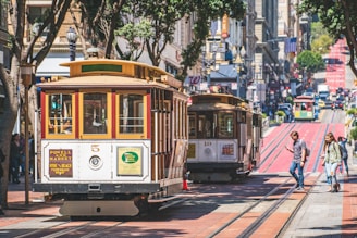 A bustling urban scene with classic cable cars traveling down a street lined with tall buildings. Pedestrians walk on the sidewalk, and the street is framed by trees. The environment suggests a lively downtown area.