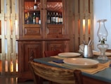 A cozy dining setting with a wooden cabinet filled with bottles, likely wine or champagne, and glassware at the top. The cabinet features intricate carvings. A table is set with plates, placemats, a metal pitcher with utensils, and a glass oil lamp. The surrounding decor includes wooden panels contributing to a warm, rustic atmosphere.