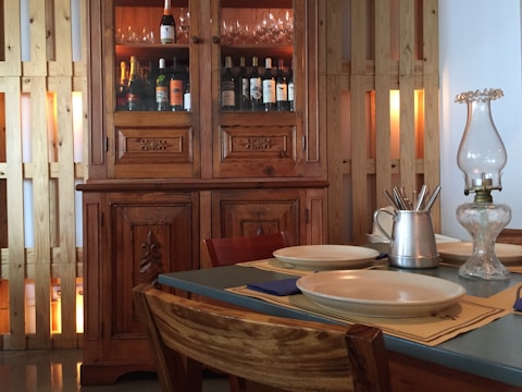 A cozy dining setting with a wooden cabinet filled with bottles, likely wine or champagne, and glassware at the top. The cabinet features intricate carvings. A table is set with plates, placemats, a metal pitcher with utensils, and a glass oil lamp. The surrounding decor includes wooden panels contributing to a warm, rustic atmosphere.