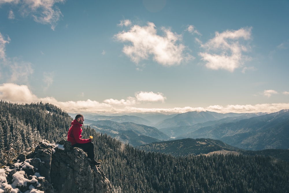 person wearing red jacket sitting on mountain peak across clouds