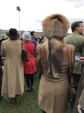 People are dressed in warm, stylish clothing, with one individual wearing a detailed fur hat. The setting appears to be outdoors with grassy ground and a cloudy sky. They are standing in a group, perhaps attending an event or gathering.