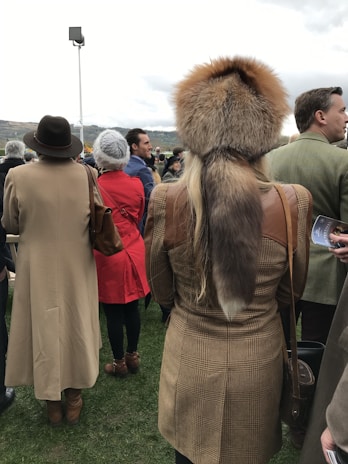 People are dressed in warm, stylish clothing, with one individual wearing a detailed fur hat. The setting appears to be outdoors with grassy ground and a cloudy sky. They are standing in a group, perhaps attending an event or gathering.