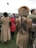 People are dressed in warm, stylish clothing, with one individual wearing a detailed fur hat. The setting appears to be outdoors with grassy ground and a cloudy sky. They are standing in a group, perhaps attending an event or gathering.