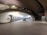 A spacious interior with a mid-century modern architectural design, featuring smooth, curved lines and an open floor. There is a classic car parked in the center, and a counter area with kiosks and digital screens on the left side. The ceiling curves elegantly, with interesting lighting fixtures casting a warm glow.