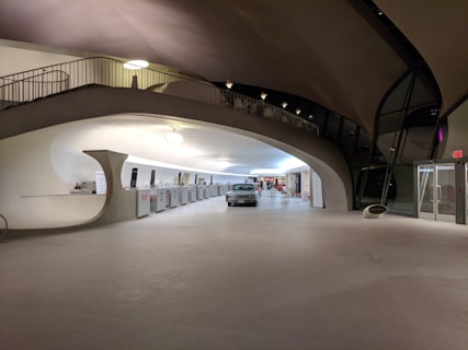 A spacious interior with a mid-century modern architectural design, featuring smooth, curved lines and an open floor. There is a classic car parked in the center, and a counter area with kiosks and digital screens on the left side. The ceiling curves elegantly, with interesting lighting fixtures casting a warm glow.