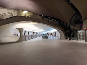 A spacious interior with a mid-century modern architectural design, featuring smooth, curved lines and an open floor. There is a classic car parked in the center, and a counter area with kiosks and digital screens on the left side. The ceiling curves elegantly, with interesting lighting fixtures casting a warm glow.