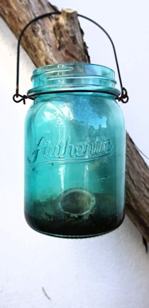 A blue glass jar with the word 'Authentic' embossed on its surface, hanging by a wire handle from a rustic wooden branch against a white wall. The jar has some dirt or debris at the bottom.
