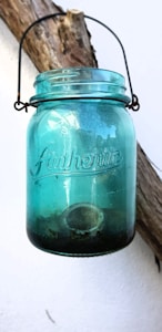 A blue glass jar with the word 'Authentic' embossed on its surface, hanging by a wire handle from a rustic wooden branch against a white wall. The jar has some dirt or debris at the bottom.