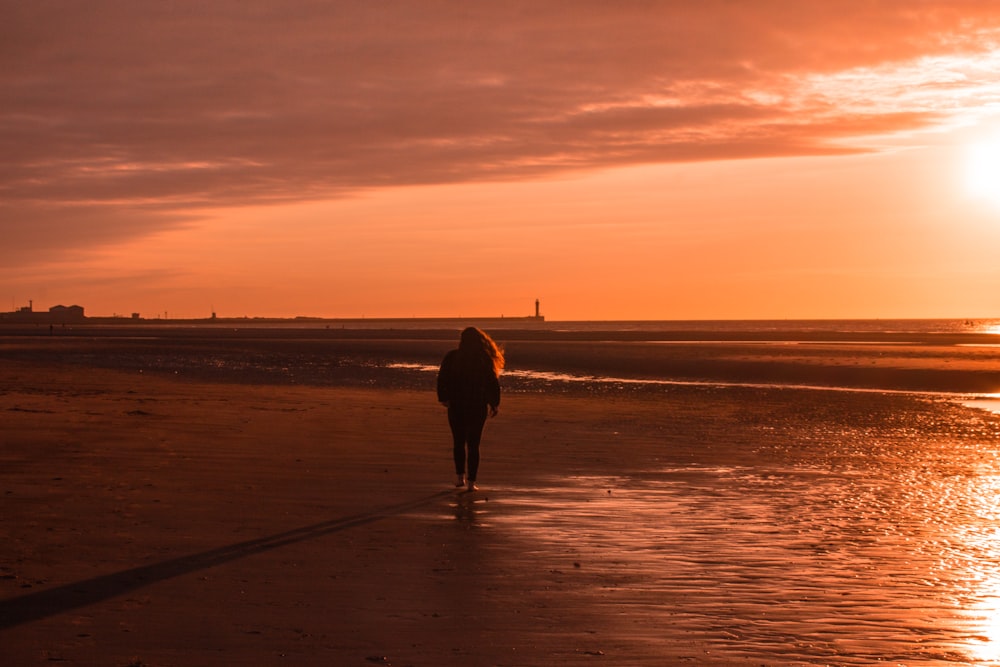 silhouette of woman walking beside seashore during sunset
