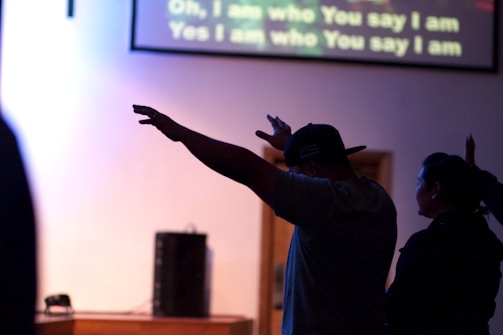 Silhouetted individuals in a dimly lit room with arms raised, possibly during a worship or concert setting. A screen in the background displays song lyrics.
