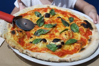 A freshly baked pizza with a crispy, slightly charred crust topped with tomato sauce, melted cheese, and fresh basil leaves. A red-handled pizza cutter is placed on the pizza, with hands visible in the background holding the plate.