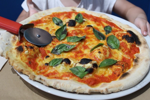 A freshly baked pizza with a crispy, slightly charred crust topped with tomato sauce, melted cheese, and fresh basil leaves. A red-handled pizza cutter is placed on the pizza, with hands visible in the background holding the plate.