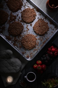 A dimly lit kitchen scene with a baking tray containing six round crunchy cookies placed on a countertop. Nearby, a small black cup holds coffee, and a lit candle casts a warm glow. Fresh berries, including strawberries, raspberries, and blackberries, are set beside the tray, alongside a gray oven mitt and a small plant.
