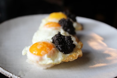 Fried eggs with bright yellow yolks are topped with a generous portion of black truffles. The eggs are neatly arranged on a white plate, with the focus on the contrasting textures and colors of the food.