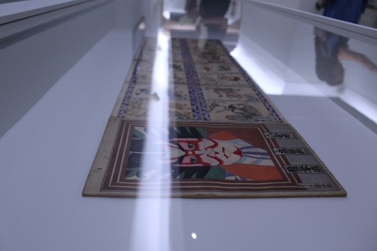 A detailed image of an artistic display in a museum or gallery setting. The artwork consists of a long scroll or panel featuring intricate illustrations and text in a showcase. The prominent image in the foreground depicts a stylized face with bold red and white patterns, reminiscent of traditional Japanese art.