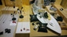 A display of various jewelry items including necklaces with red pendants, stylish bracelets, and earrings on a wooden surface. The centerpiece is a white tray holding a silver fish can, lemon slices, and black cushion pads with jewelry. A small blackboard with written text is visible in the background.