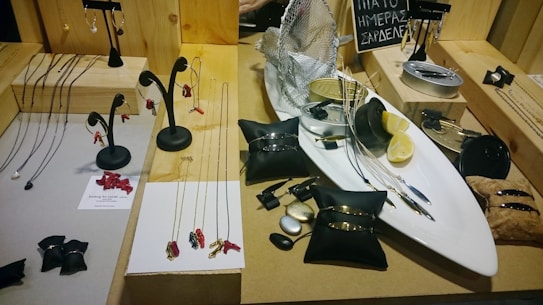 A display of various jewelry items including necklaces with red pendants, stylish bracelets, and earrings on a wooden surface. The centerpiece is a white tray holding a silver fish can, lemon slices, and black cushion pads with jewelry. A small blackboard with written text is visible in the background.