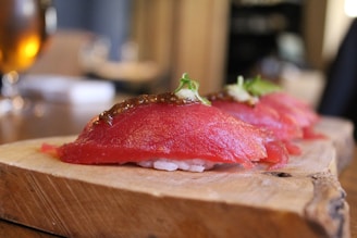 Three pieces of sushi placed on a wooden serving board, featuring a slice of raw red fish on top of rice, garnished with herbs and a dollop of sauce.
