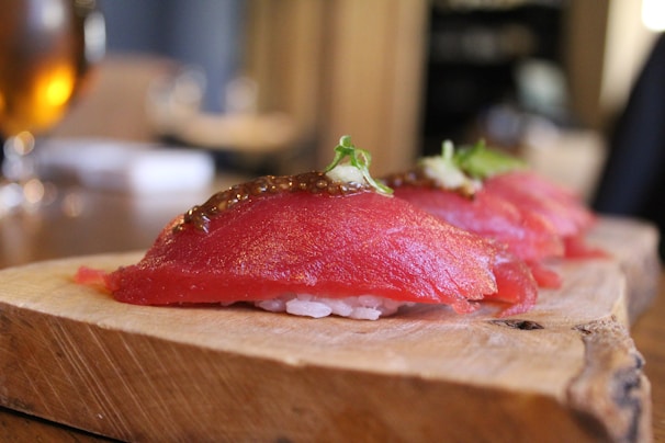 Three pieces of sushi placed on a wooden serving board, featuring a slice of raw red fish on top of rice, garnished with herbs and a dollop of sauce.