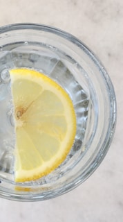 A glass of water with a slice of lemon, showcasing clear liquid and the lemon's bright yellow peel. Small bubbles are visible on the glass surface.