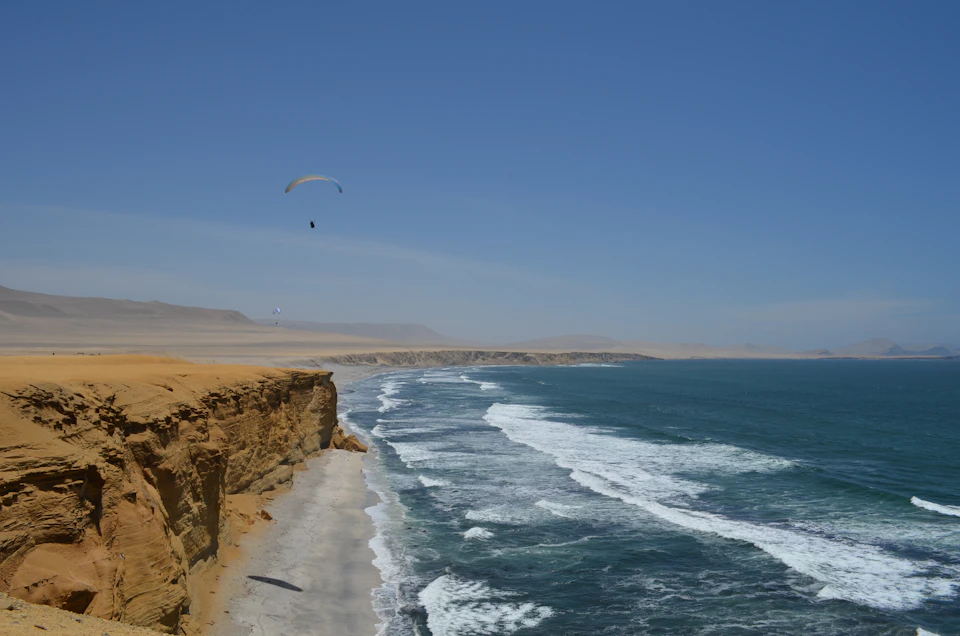 The image depicts a serene beach scene with a cliff on one side and a body of water on the other, both covered in white foam from crashing waves. A person is seen flying a kite high above the ocean, adding a dynamic element to the otherwise tranquil setting. The sky is clear blue, providing a beautiful backdrop for the scene.
