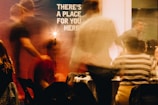 Blurred figures are seen in a warmly lit indoor setting, suggesting a social gathering. A prominent sign in the background reads 'There's a place for you here.' People appear to be in motion, creating a dynamic and lively atmosphere.
