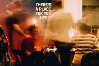 Blurred figures are seen in a warmly lit indoor setting, suggesting a social gathering. A prominent sign in the background reads 'There's a place for you here.' People appear to be in motion, creating a dynamic and lively atmosphere.