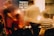 Blurred figures are seen in a warmly lit indoor setting, suggesting a social gathering. A prominent sign in the background reads 'There's a place for you here.' People appear to be in motion, creating a dynamic and lively atmosphere.