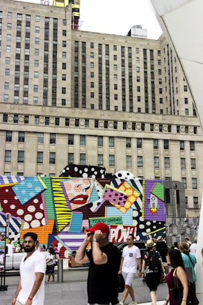 A large building serves as a backdrop for a vibrant, colorful mural featuring pop art elements, including bold patterns and stylized graphics of a face with red lips. People walk along the street in front of the mural, some appearing to be tourists capturing the scene. The mural and the urban surroundings create a lively atmosphere.