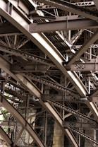 Intricate metal beams and structures form an elaborate framework, showcasing industrial design and engineering. The complex web of girders features rivets and cross-bracing against a backdrop of masonry.