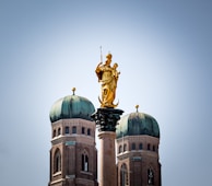 A golden statue stands prominently on a tall column with two rounded domed towers in the background against a clear blue sky.