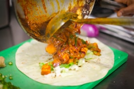 A filling of colorful, cooked vegetables is being poured from a pan onto a flat tortilla placed on a green cutting board. The vegetables are covered in a rich, spicy-looking sauce.