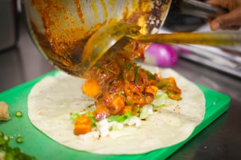 A filling of colorful, cooked vegetables is being poured from a pan onto a flat tortilla placed on a green cutting board. The vegetables are covered in a rich, spicy-looking sauce.