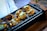 A tray of toasted bread slices topped with a mixture of melted cheese and seasoned vegetables. The bread is presented on a black grill pan with a brown wooden handle, alongside a pair of large metal tongs.
