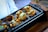 A tray of toasted bread slices topped with a mixture of melted cheese and seasoned vegetables. The bread is presented on a black grill pan with a brown wooden handle, alongside a pair of large metal tongs.