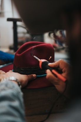 A person is using a tool to engrave or burn a design into a red felt hat. The hat is held on a workbench, and the person's hands are visible, indicating careful craftsmanship.