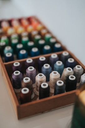 A wooden box filled with multiple spools of thread organized in rows, featuring a variety of colors including shades of blue, green, purple, brown, and orange. The focus is on the front row, while the background is slightly blurred.