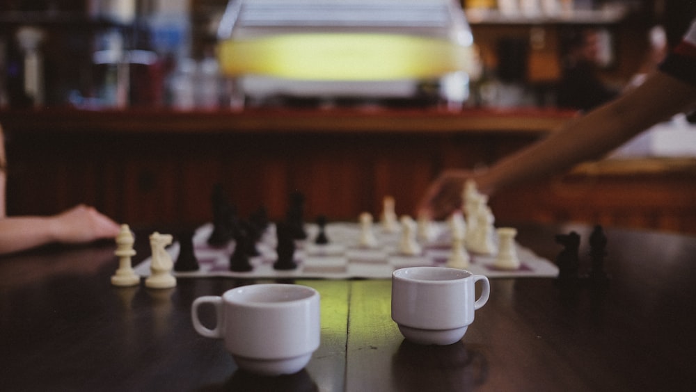 zwei weiße Kaffeetassen auf dem Tisch neben zwei Leuten, die Schach spielen