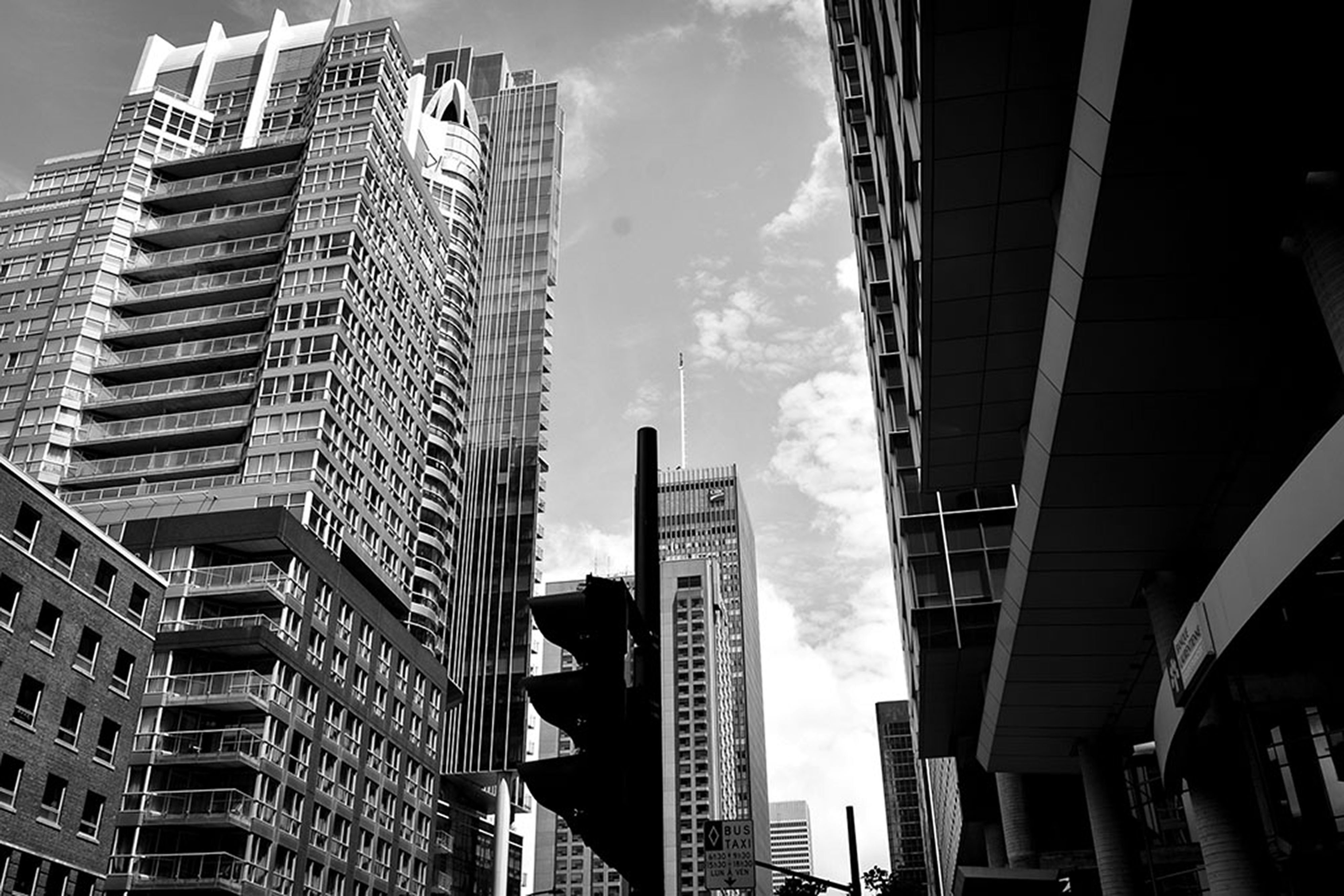 A black and white cityscape featuring tall skyscrapers with modern architectural designs. The structures vary in height, creating a dynamic skyline against a partly cloudy sky. A prominent traffic signal silhouette is visible in the foreground, adding an urban element.