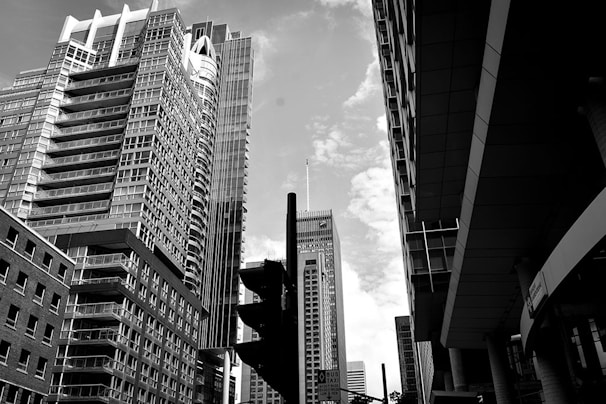 A black and white cityscape featuring tall skyscrapers with modern architectural designs. The structures vary in height, creating a dynamic skyline against a partly cloudy sky. A prominent traffic signal silhouette is visible in the foreground, adding an urban element.