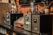 A row of vintage audio amplifiers is displayed on a dark wooden shelf, with metal dials and glass vacuum tubes prominently visible. The background includes a rustic brick wall and a decorative ceramic vase with dried branches and flowers.
