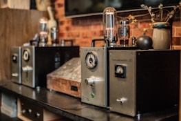 A row of vintage audio amplifiers is displayed on a dark wooden shelf, with metal dials and glass vacuum tubes prominently visible. The background includes a rustic brick wall and a decorative ceramic vase with dried branches and flowers.
