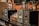 A row of vintage audio amplifiers is displayed on a dark wooden shelf, with metal dials and glass vacuum tubes prominently visible. The background includes a rustic brick wall and a decorative ceramic vase with dried branches and flowers.