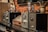 A row of vintage audio amplifiers is displayed on a dark wooden shelf, with metal dials and glass vacuum tubes prominently visible. The background includes a rustic brick wall and a decorative ceramic vase with dried branches and flowers.