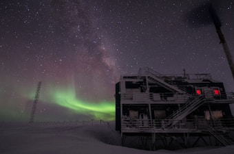 A landscape featuring a night sky illuminated by the Milky Way and a vivid green aurora borealis. The scene includes a large building, possibly a scientific research station, with staircases and bright red lights in the windows. Snow covers the ground and a tall tower structure is present.
