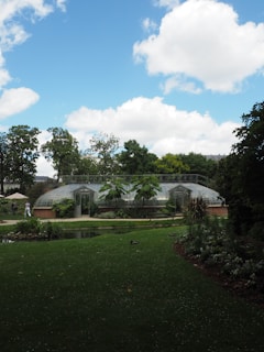A park scene with a lush green lawn and trees under a bright blue sky with fluffy white clouds. A greenhouse with glass panels is centrally located, surrounded by various plants and greenery. A small pond is visible in the foreground, with a person walking in the background.