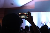 A person holds up a smartphone to record an event or performance. The scene is dimly lit, with a bright stage in the background where individuals appear to be standing or presenting. The smartphone screen shows a clearer view of the stage, capturing both the stage setting and the people on it.