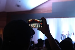 A person holds up a smartphone to record an event or performance. The scene is dimly lit, with a bright stage in the background where individuals appear to be standing or presenting. The smartphone screen shows a clearer view of the stage, capturing both the stage setting and the people on it.