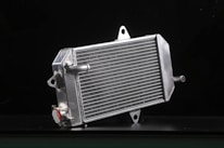 A metallic car radiator is placed against a dark background, highlighting its shiny silver surface and intricate grid of cooling fins. The radiator has several metal connectors protruding from its sides, and a cap with a caution label is visible in the lower area.