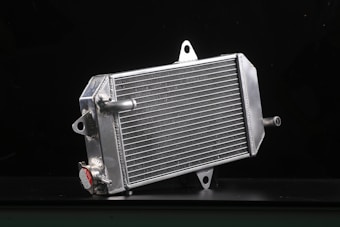 A metallic car radiator is placed against a dark background, highlighting its shiny silver surface and intricate grid of cooling fins. The radiator has several metal connectors protruding from its sides, and a cap with a caution label is visible in the lower area.